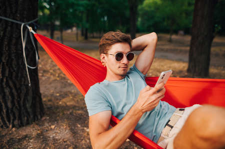 Handsome young man in casual clothes resting in the woods lying in a hammock on a tree and using a smartphone with a serious face.の写真素材