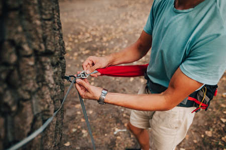 A close-up photo of the hands of a male tourist ties the hammock slings to a tree. Creating a campsite in the woods. Ties a hammock to a tree for campingの写真素材
