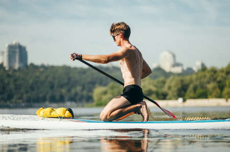 Young male athlete with muscular torso trains on sup board at sea, rowing with oar. Active recreation on the water.の写真素材
