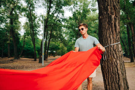 Handsome young man touches a red tourist hammock to a tree in the forest with a serious face.の写真素材