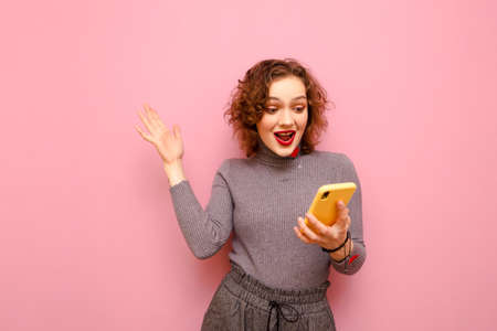 Portrait of shocked girl with curly hair stands on pink background, looks at smartphone screen with...
