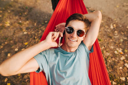 Positive guy in sunglasses lies in a hammock on a tree in the woods, talking on the phone and looking at the camera with a smile on his face.の写真素材
