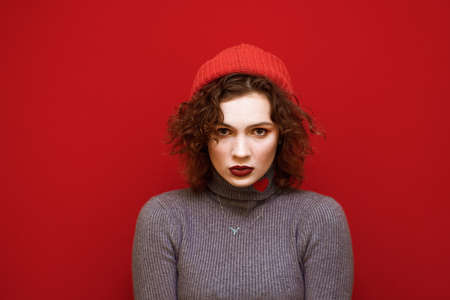 Closeup portrait of a curly-haired lady in a gray sweater with a hat on her head looking into the camera with a serious face. Serious young model in hat and freckles isolvoan on red background.の写真素材