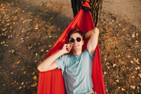 Portrait of a happy young man in sun colari and t-shirt lying in a red hammock on a tree in the...