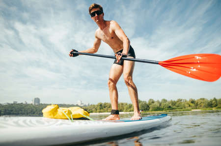 Muscular young man paddles on a sup board by the sea, and looks at the camera. Portrait of a sporty young man training on a rowing board.の写真素材