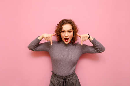 Astonished curly girl in a sweater stands on a pink background, looks into the camera and shows her thumbs down on copy space. Shocked attractive lady showing thumbs down to empty space.の写真素材