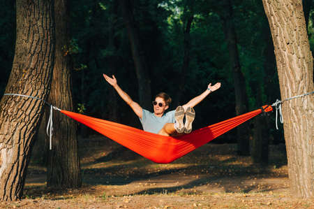 Portrait of a joyful young man in sunglasses sitting on a hammock in the park and swaying with a smile on his face and arms raised.の写真素材