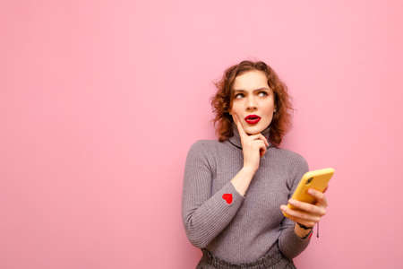 Portrait of pensive girl in gray sweater stands on pink background with smartphone in hand and looks...