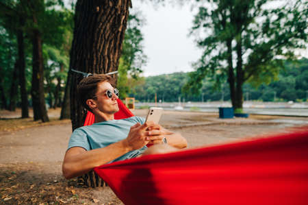 Young handsome man resting in the park lying on a hammock with a smartphone in his hands, looking away with a serious face.の写真素材