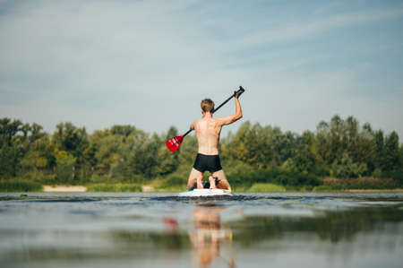 Back of a young sports man is actively rowing on the river on a sup board. Active recreation on the water with a rowing board.の写真素材