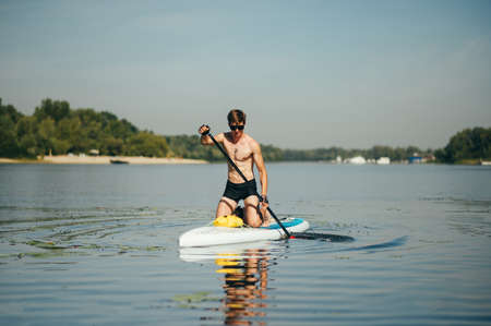 Athletic handsome man paddles on a sup board on the river. Training on a rowing board on a sunny...