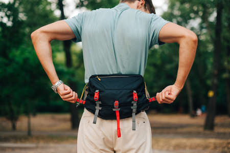 Man in casual clothes stands in the woods during the day and fastens fastex on a waist bag. Tourist...