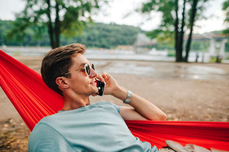 Handsome young man talking on the phone in the park on a hammock against the backdrop of a beautiful landscape, looking away at copy space and smiling.の写真素材