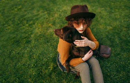 Fashionable girl in vintage clothes sits with a dog on her hands on a green lawn, hugging a pet....