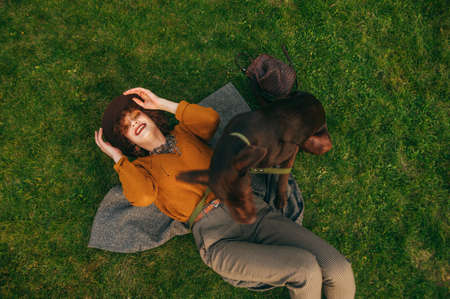 Smiling girl in vintage clothes and hat playing with dog on lawn, top view. Cheerful owner relaxing...