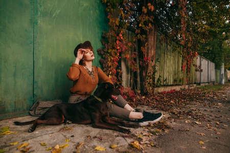 Fashionable lady sits outside in autumn day near country house and looks away against the backdrop of beautiful autumn countryside landscape. Copy spaceの写真素材