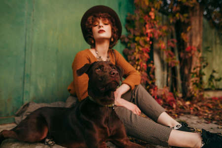 Portrait of a beautiful dog sitting with the owner on the ground in the village on a background of autumn yard, looking away, girl blurred on the background. Pet and fashionable owner autumn photo.の写真素材