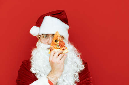 Closeup portrait of Santa Claus holding a slice of pizza in front of his eye and looking into the camera with a serious face isolated on red background. Copy spaceの写真素材
