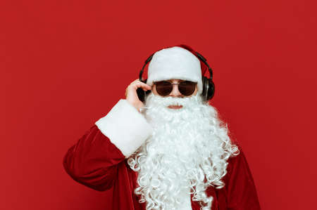 Studio closeup portrait of cool Santa Claus in sunglasses listening to music in headphones and looking at camera on red background. The cool modern Santa loves to listen to music. Xmas. Copyspaceの写真素材