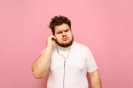 Funny bearded young fat man with displeased face listening to music in headphones and looking into camera. Cheerful guy with overweight and curly hair in headphones isolated on pink background.の写真素材