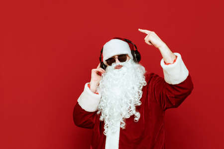 Studio portrait of cheerful Santa Claus in sunglasses listening to music in headphones and having fun on red background. Santa Claus partyman with headphones isolated on red background. X-masの写真素材