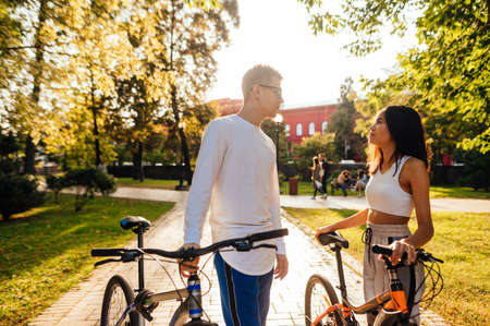Beautiful young couple on bicycles stands in the park in autumn season and communicates on a background of sunset. Beautiful woman and man on a bike ride.の写真素材