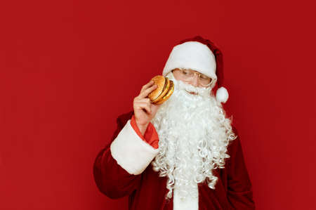 Portrait of serious Santa Claus in glasses and white beard isolated on red background with burger in hand, looking aside for fast food with serious face. Funny Santa Claus eats fast food. Christmasの写真素材