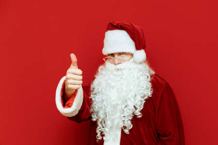 American Santa Claus stands on a red background, shows thumb up and looks at the camera with a serious face. Portrait of Santa with elongated finger up, isolated. Santee likes it. Christmas.X-masの写真素材
