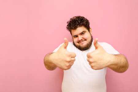 Cheerful bearded fat man in white t-shirt isolated on pink background, looks into camera and shows thumbs up. Positive guy with overweight and curly hair shows gesture like.の写真素材