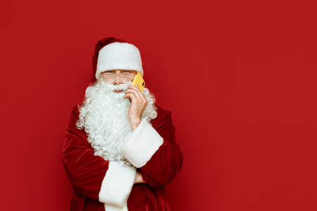 Portrait of a funny santa stands on a red background and talks on the phone with a serious face. Santa is calling on smartphone. Isolated. Christmas concept. Copyspaceの写真素材