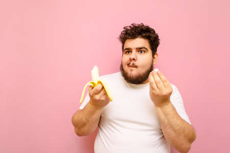 Funny curly fat man with a beard eats a banana on a pink background, looks away with a funny face. Positive young overweight man in white t-shirt isolated on pink background eating banana.の写真素材