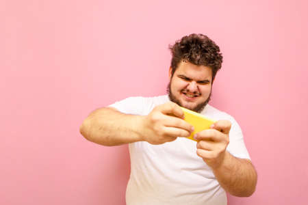 Emotional fat man in white t-shirt playing mobile online games on smartphone with expressive face. Concentrated overweight guy playing mobile games on a smartphone.の写真素材