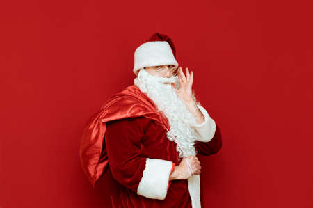 Portrait of a man in a suit of Santa Claus stands on a red background with a bag of gifts on his...