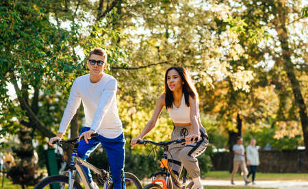 Stylish beautiful couple rides bicycles in the autumn park. Young stylish people walking on a bicycle through the city park in the autumn season.の写真素材