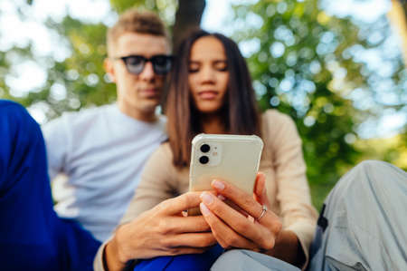 Couple sitting in the park and using the Internet on a smartphone, looking at the screen. Focus on...