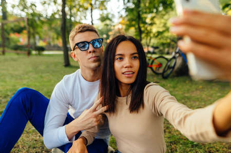 Beautiful young couple sitting on the lawn in the park and taking a selfie on a smartphone. Woman...