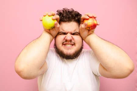 Close up portrait of emotional fat guy with apples in his hands, looking dissatisfied into the camera and yelling. Hungry fat man with apples in hands isolated on pink background.の写真素材