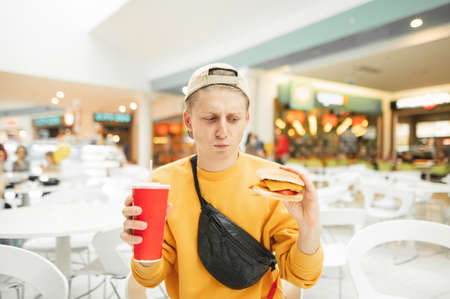 Funny young man stands with a burger and a glass of cola in his hand and looks upset at a fast food. Guy the student in the cap is forced to eat fast food, he is dissatisfied. Junk food concept.の写真素材