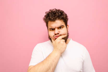 Funny fat man in white t-shirt looks into camera with pensive serious face on pink background, isolated. Funny pensive guy with overweight, beard and curly hair, isolated.の写真素材