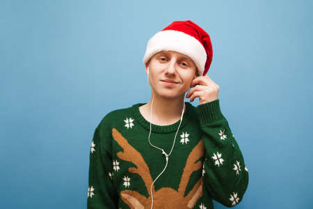 Closeup portrait of funny guy in christmas sweater and santa hat on blue background, looking into...
