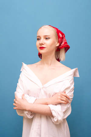 Attractive lady in white overseer shirt and red handkerchief isolated on blue background, looking away with a smile on her face. Fashionable photo of a girl in a headscarf and a man's shirt.の写真素材