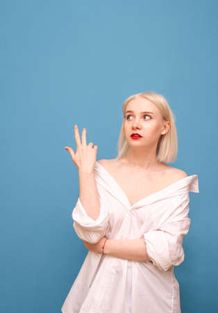 Vertical portrait of an attractive lady in a man's shirt stands on a blue background with a bewildered face, looking to the side and showing thumbs up in a blank space. Copy spaceの写真素材