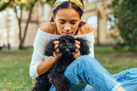 Close portrait of beautiful owner and black little dog hugging in park sitting on grass. The cute owner hugs a toy poodle dog.の写真素材