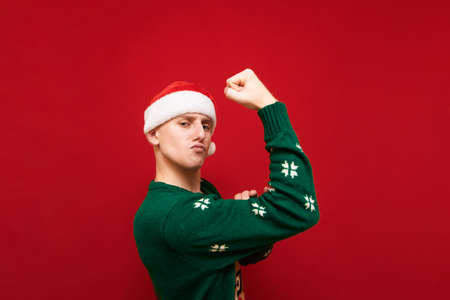 Funny guy with serious face stands on red background in christmas clothes and shows biceps. Funny guy in santa hat showing biceps. Copy spaceの写真素材