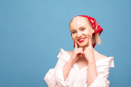 Smiling girl in white over-sized shirt and red headscarf, isolated on blue background, looks into camera with happy face. Portrait of attractive peasant girl on blue background with smile on her face.の写真素材