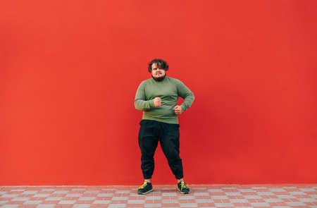Funny young overweight man dances on a red background and looks away with a smile on his face. Full-length portrait of a dancing fat man.の写真素材