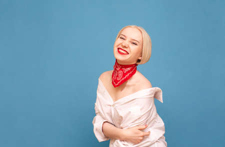 Happy sexy lady in red scarf around her neck and men's shirt isolated on blue background, looks into the camera with a smile on her face. Joyful lady in white dress shirt poses at camera.の写真素材