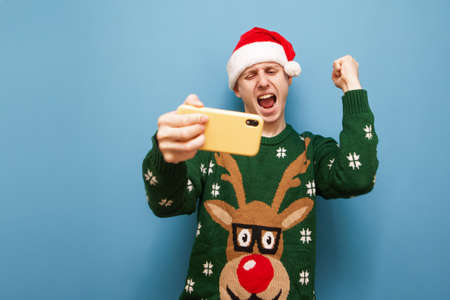 Emotional guy gamer in Christmas clothes playing mobile games on a blue background, rejoices with a...
