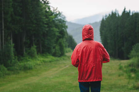 Back view of a tourist in a vivid red waterproof coat wearing a hood in a rainy weather in the...