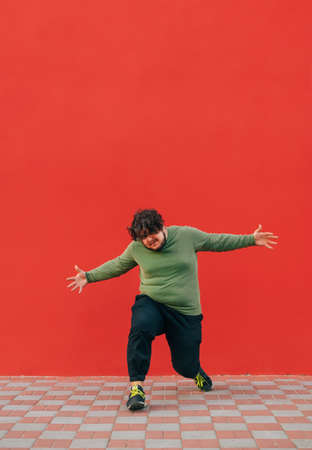 Fat curly young man in stylish street clothes dancing hip hop on a red wall background. Vertical.